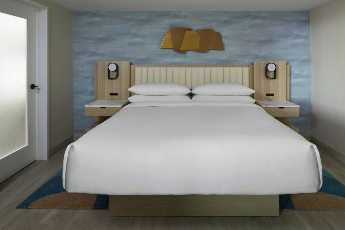 Una habitación de hotel con una cama grande, dos mesitas de noche, un cabecero con lámparas de pared y una puerta.
