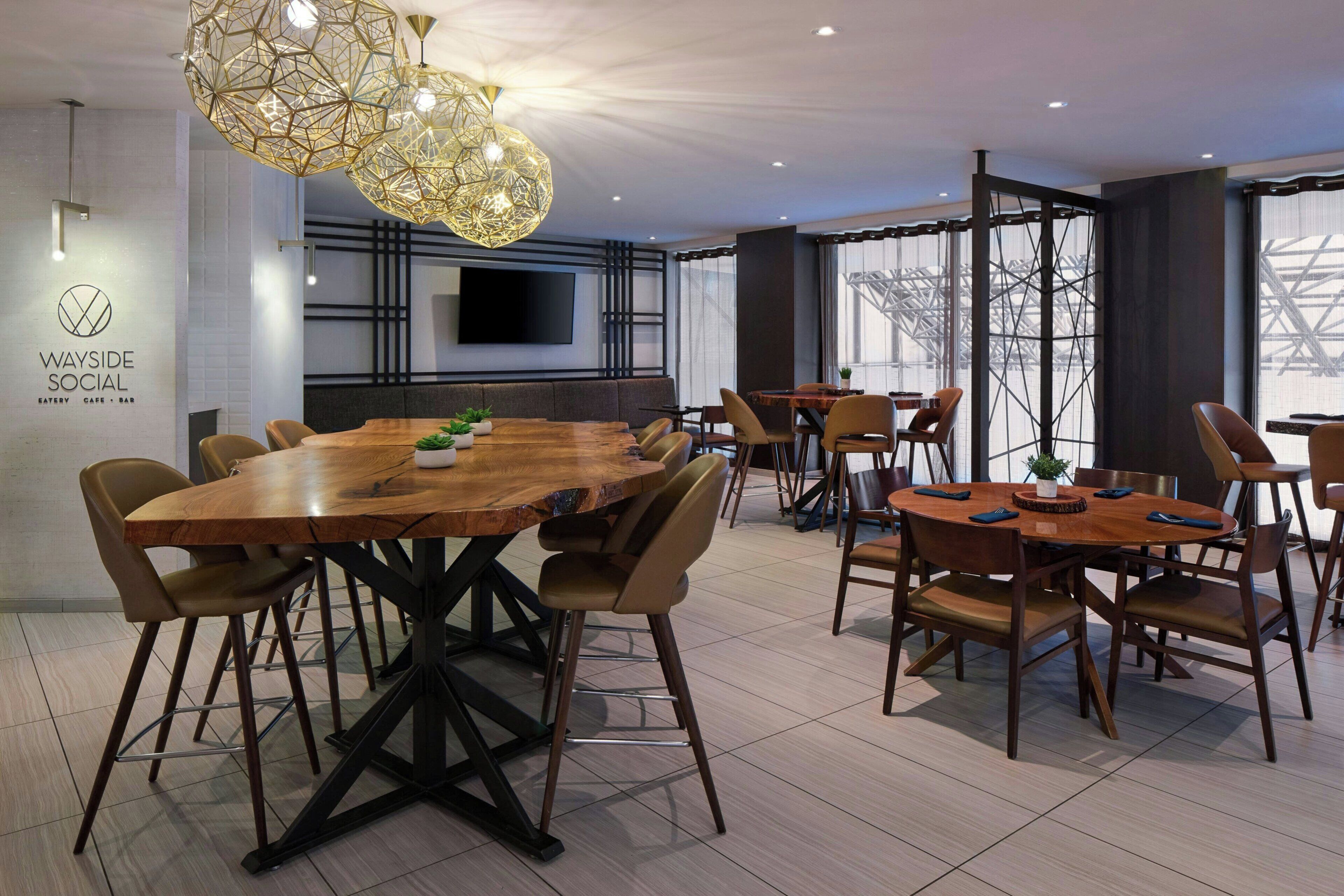 A modern dining area with a large wooden table, multiple smaller tables, and comfortable chairs. There is a large, geometric chandelier hanging from the ceiling, and a flat-screen TV mounted on the wall.