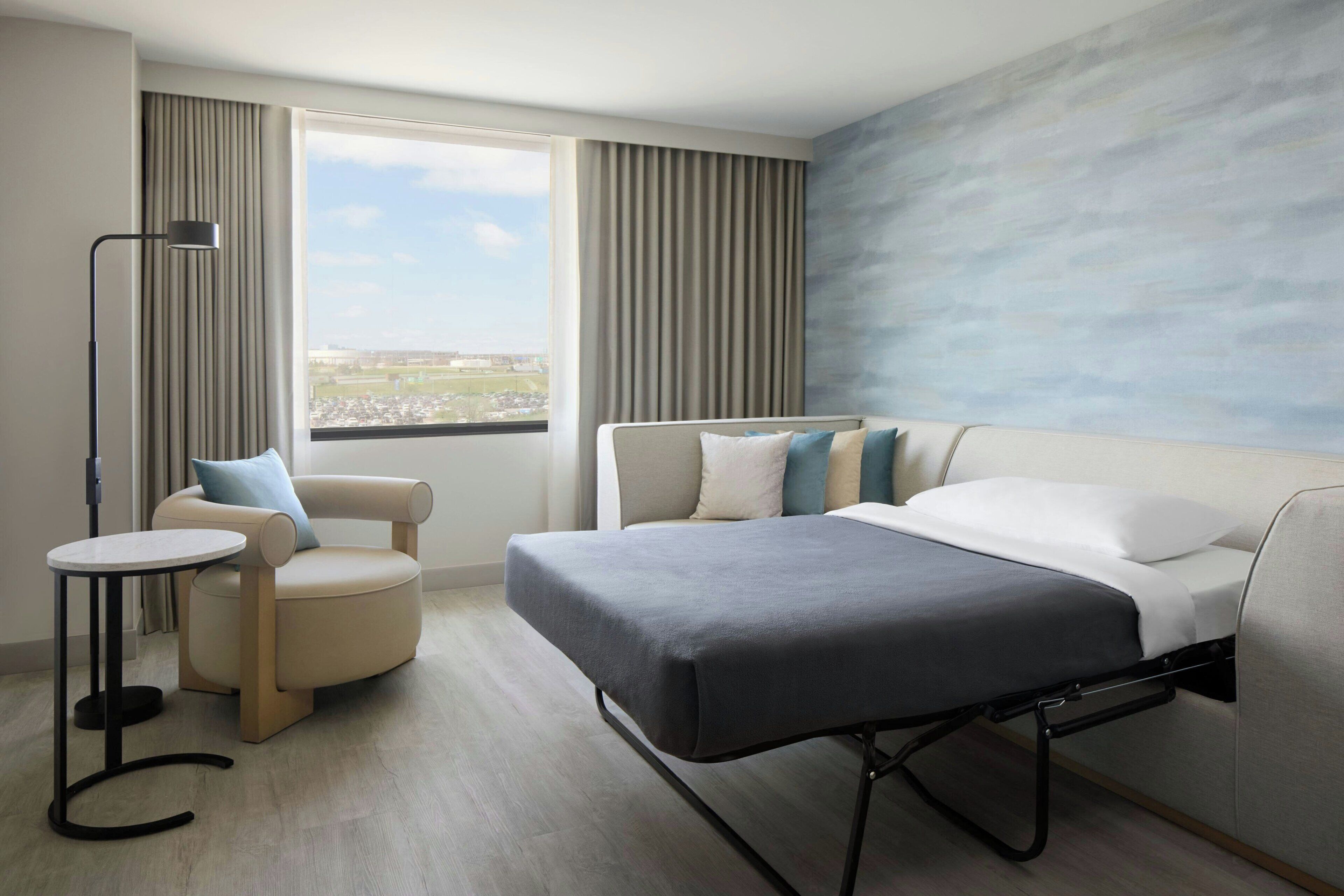 A hotel room with a bed, a chair, a small table, a window with curtains, and a city view.