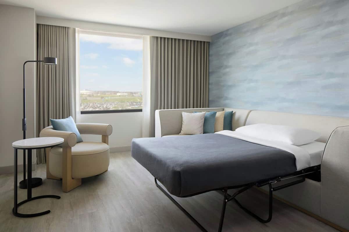 Une chambre d’hôtel avec un lit, une chaise, une petite table, une fenêtre avec des rideaux et une vue sur la ville.
