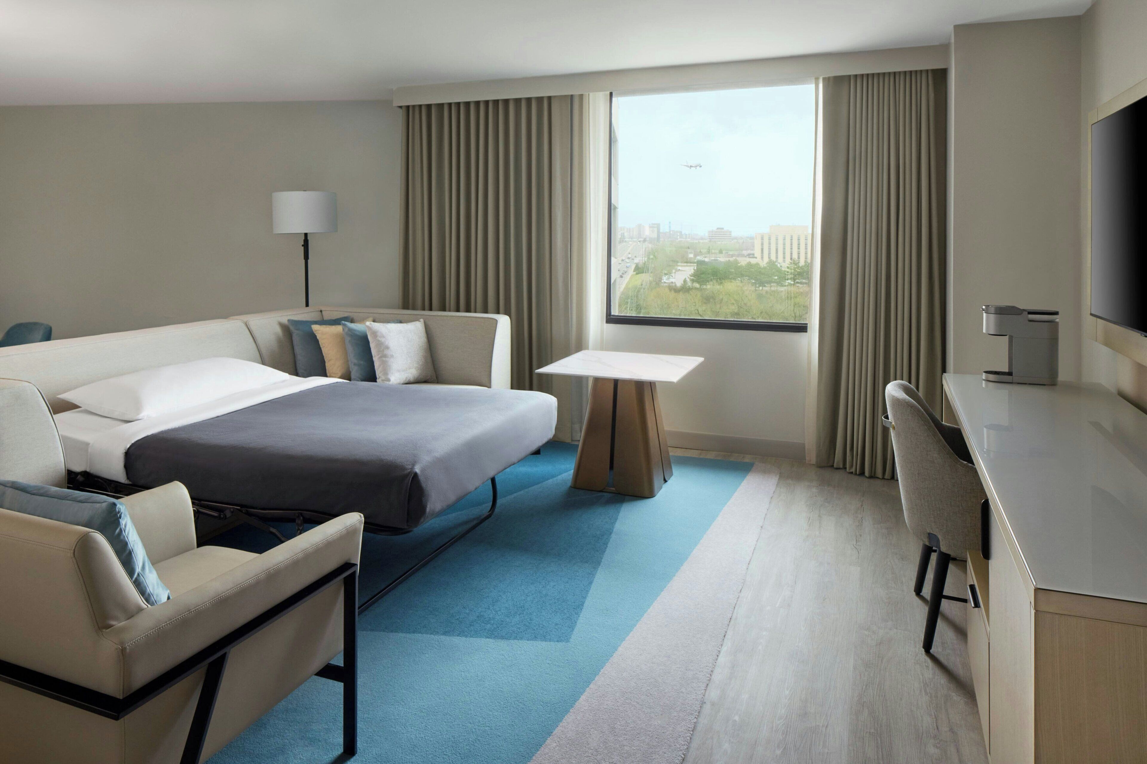 A hotel room with a bed, a desk, a chair, a TV, and a window with a view.