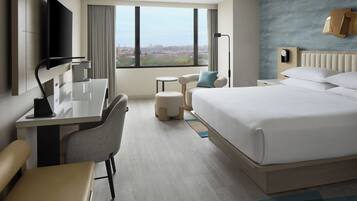 A hotel room with a bed, a desk, a chair, a small table, and a view of the city through large windows.
