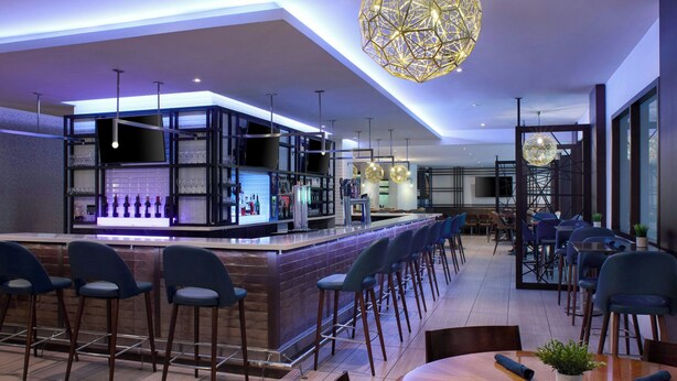 Un área de bar moderna con barra, taburetes y un espacio de estar con mesas y sillas.
