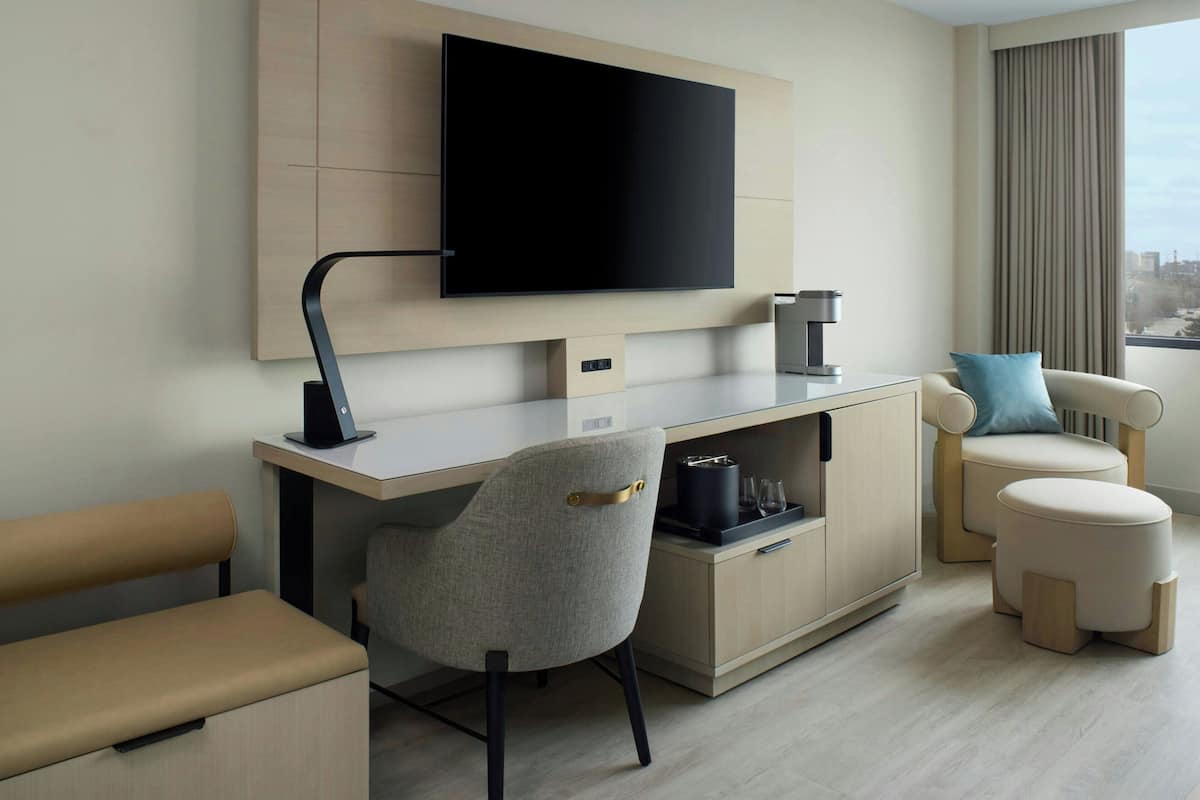 A modern hotel room with a flat-screen TV mounted on the wall, a desk with a chair, a sofa, and a small ottoman.