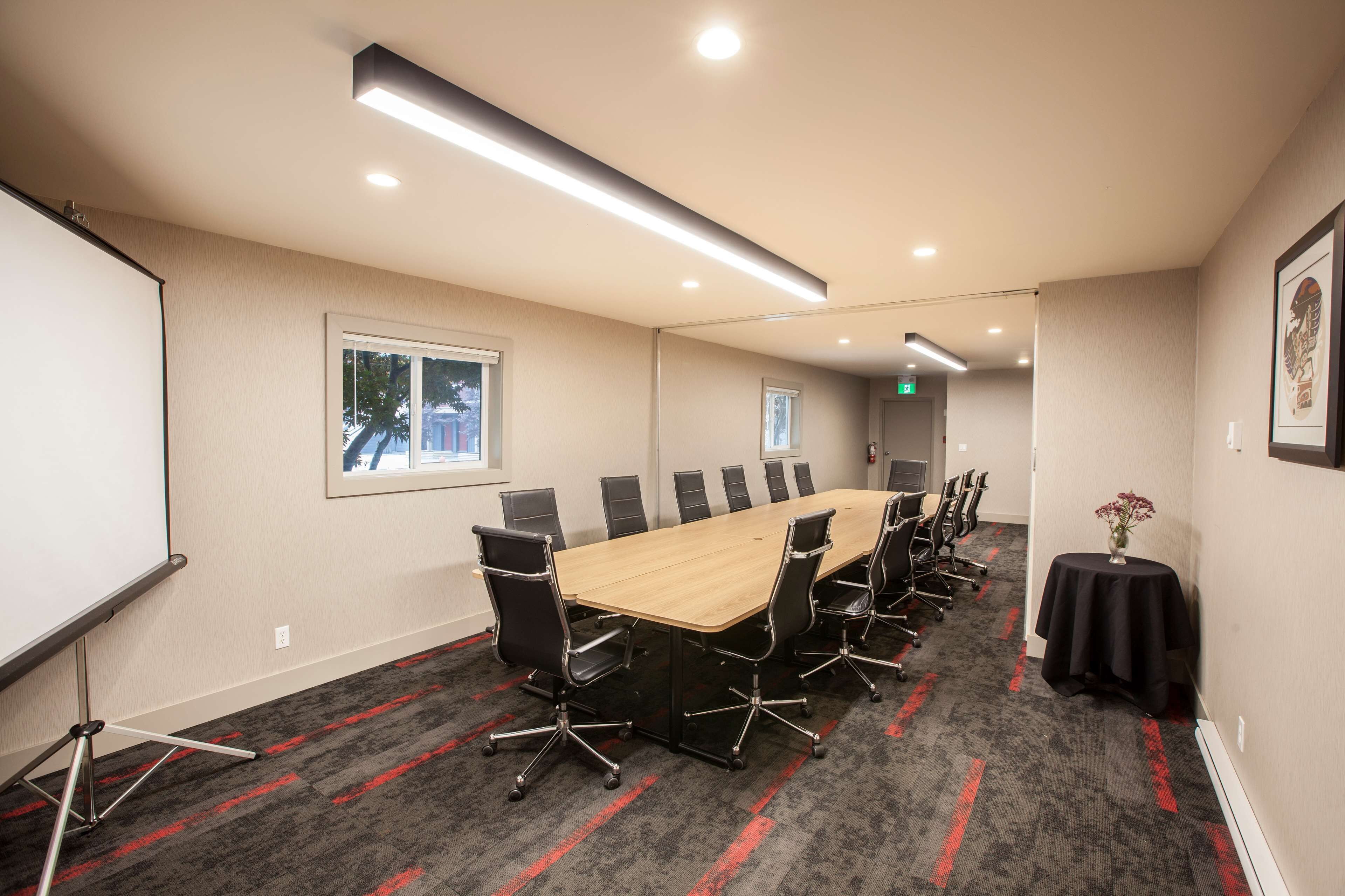 A conference room with a long table, black chairs, a projector screen, and a small table with a vase.