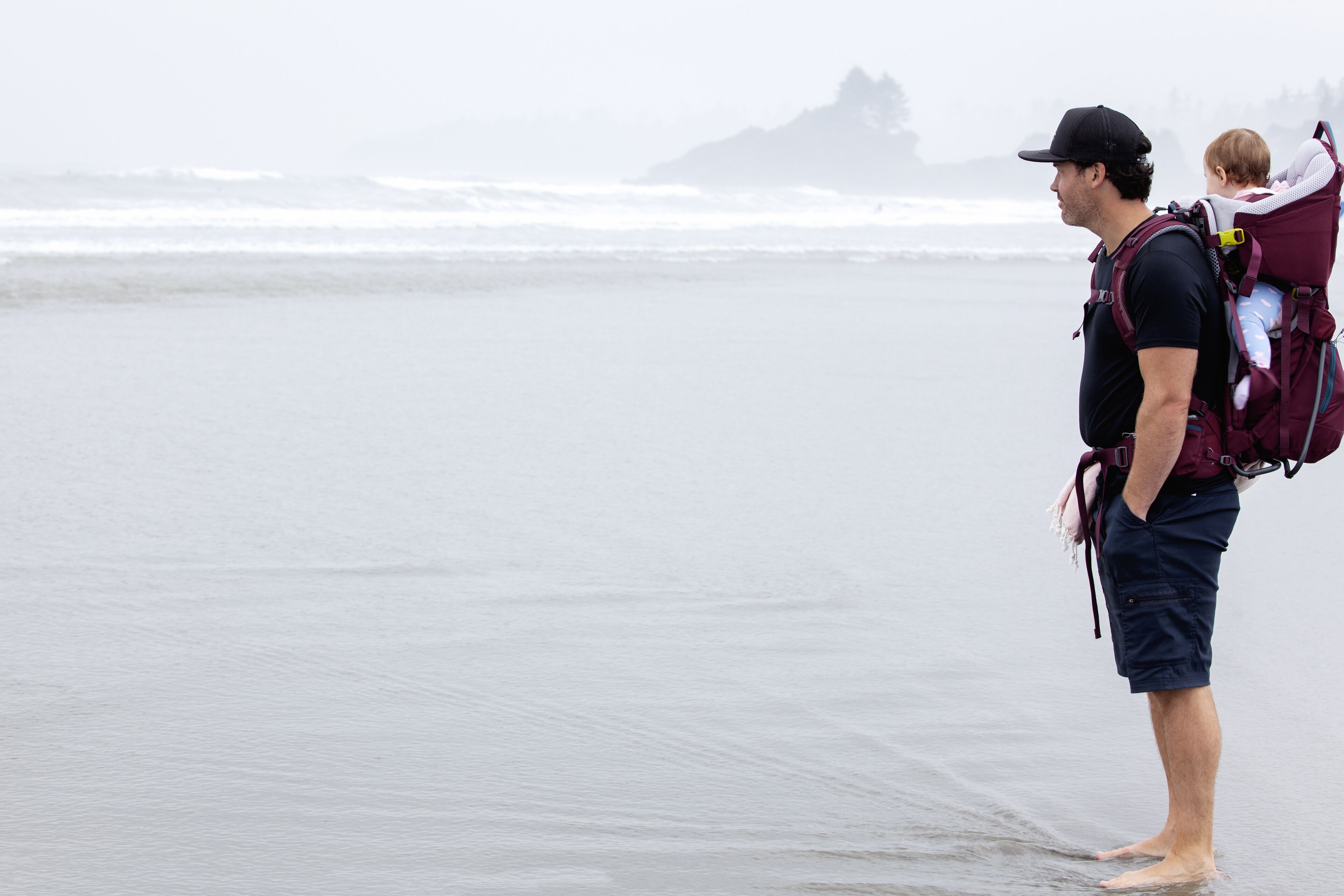 A person standing on a beach with a child in a carrier on their back.