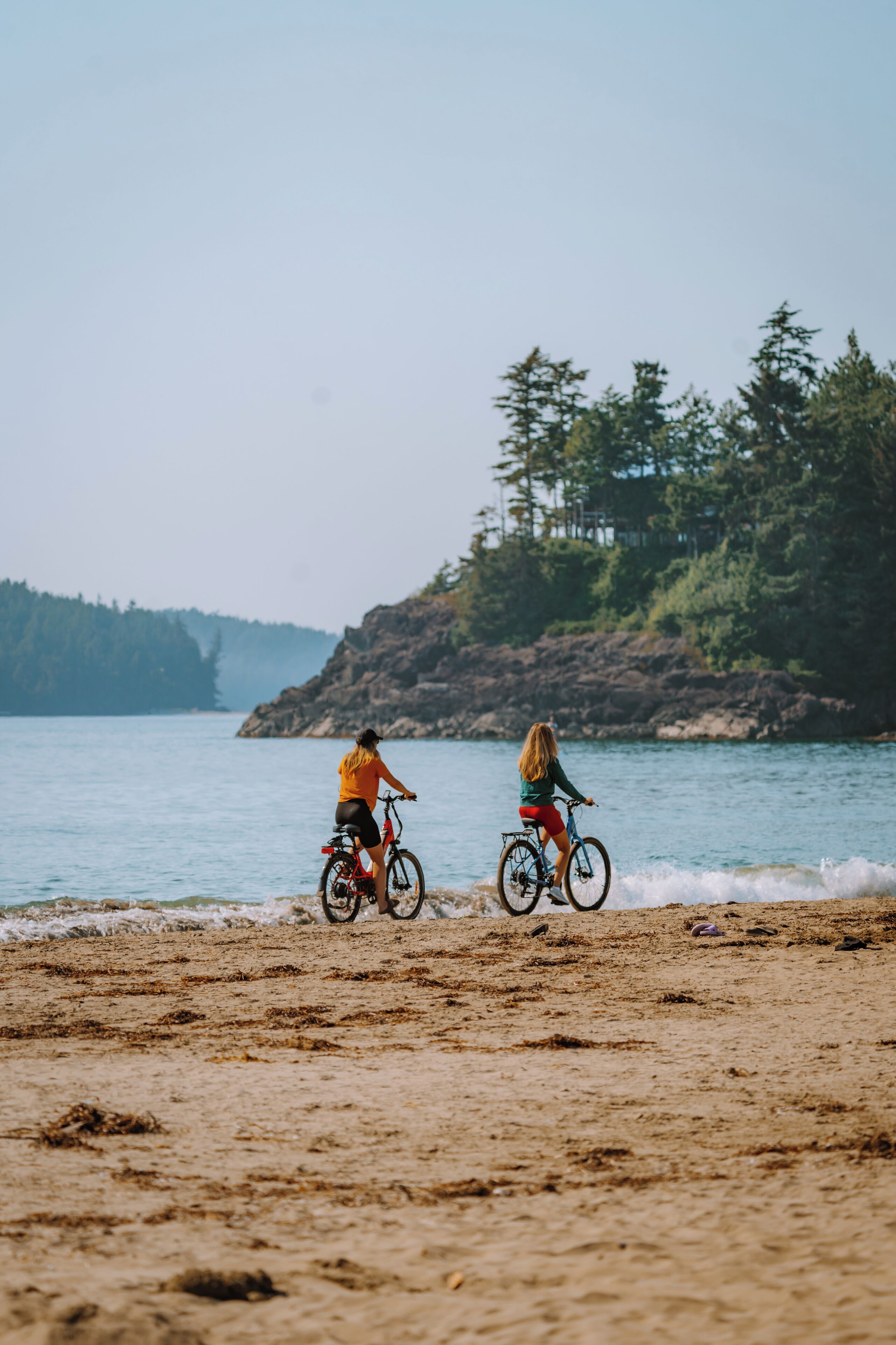 Dos personas paseando en bicicleta sobre una playa arenosa con costa rocosa y árboles al fondo.