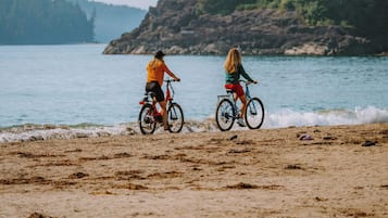 Two people riding bicycles on a sandy beach with a rocky shoreline and trees in the background.