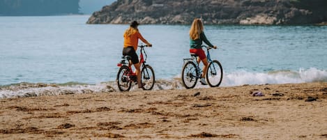 Dos personas paseando en bicicleta sobre una playa arenosa con costa rocosa y árboles al fondo.