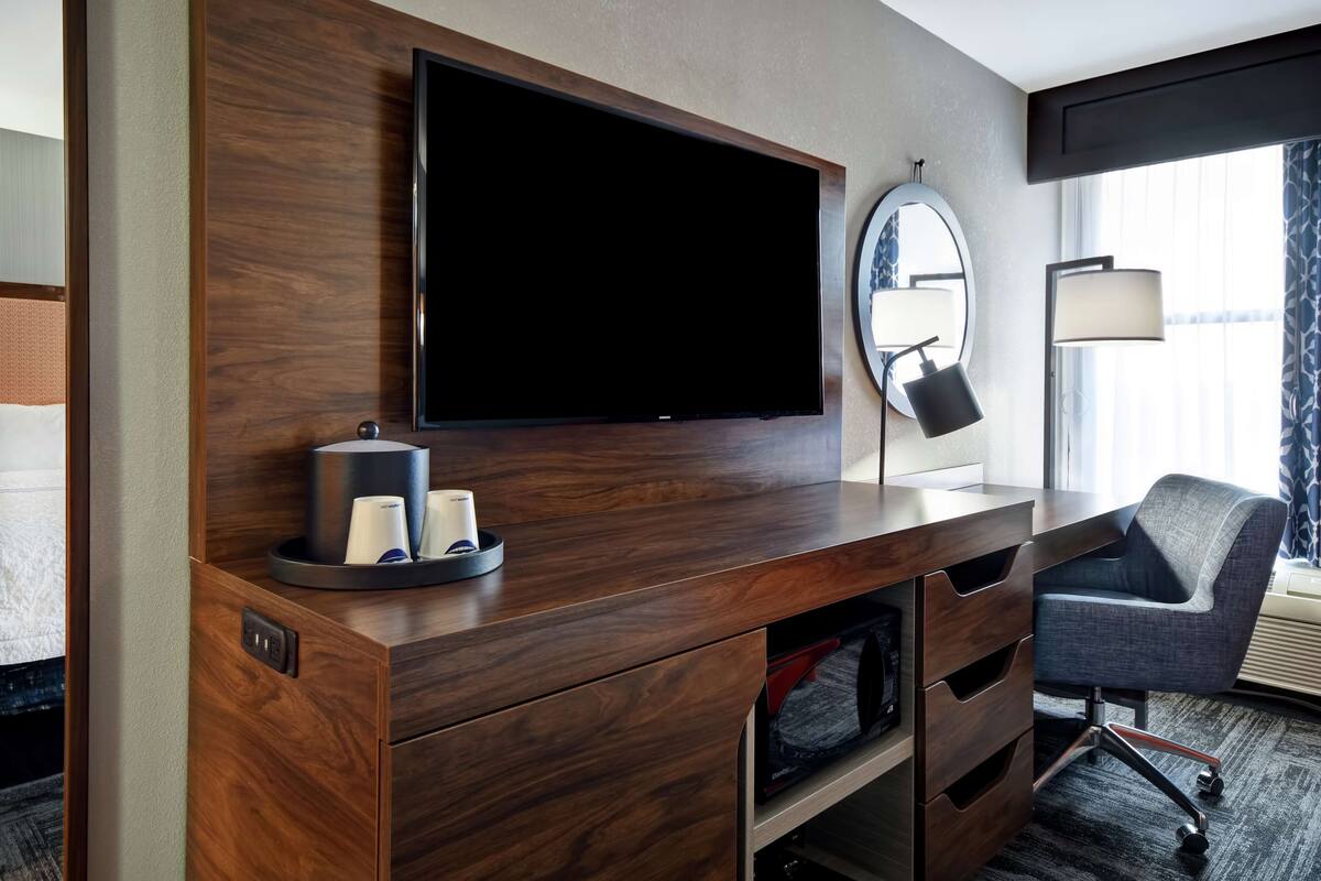 A hotel room with a wooden desk, a flat-screen TV, a mirror, a lamp, and a chair.