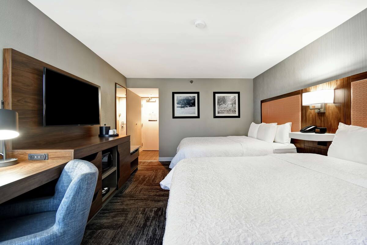 A hotel room with two beds, a desk with a flat-screen TV, a chair, and a bedside table with a lamp.