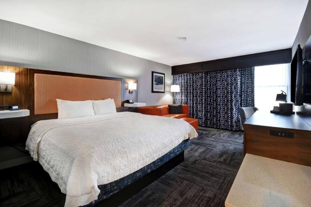 A hotel room with a large bed, a desk, a chair, a TV, and a window with patterned curtains.