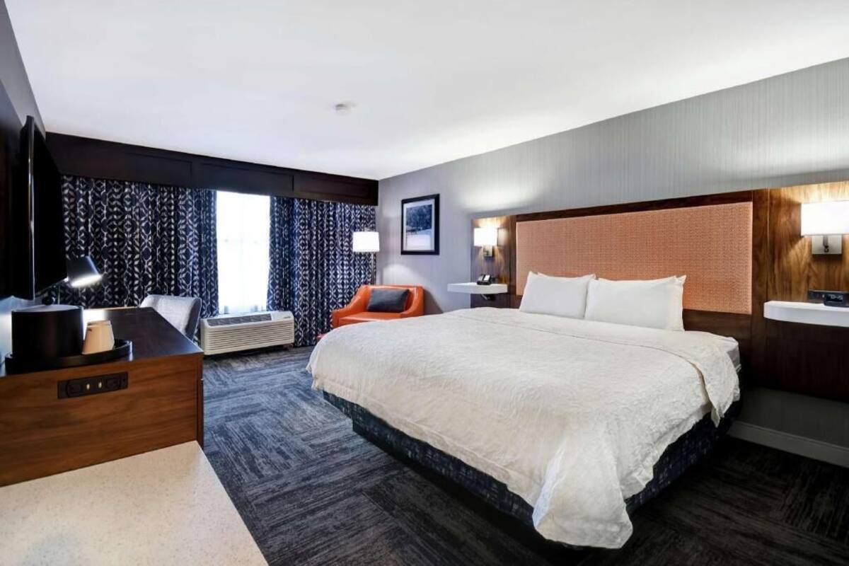 A hotel room with a large bed, a desk with a lamp, a chair, a bedside table, and a window with patterned curtains.