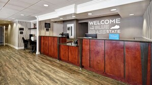 A reception desk with a "Welcome to the Blue Grass State" sign, a wooden reception desk, and a "Hilton Honors" sign. - Hampton Inn I-75 Lexington/Hamburg Area (Lexington)