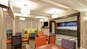 A hotel dining area with tables and chairs, a television, and a kitchen in the background. - Hampton Inn I-75 Lexington/Hamburg Area (Lexington)