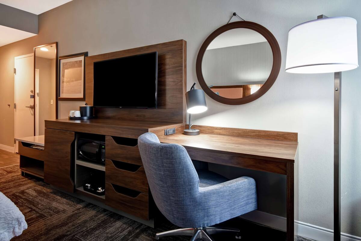 A modern hotel room with a wooden desk, a flat-screen TV, a round mirror, and a blue chair.
