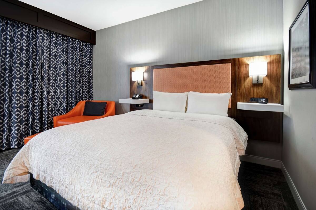 A hotel room with a large bed, two bedside tables with lamps, a chair, and a patterned curtain.