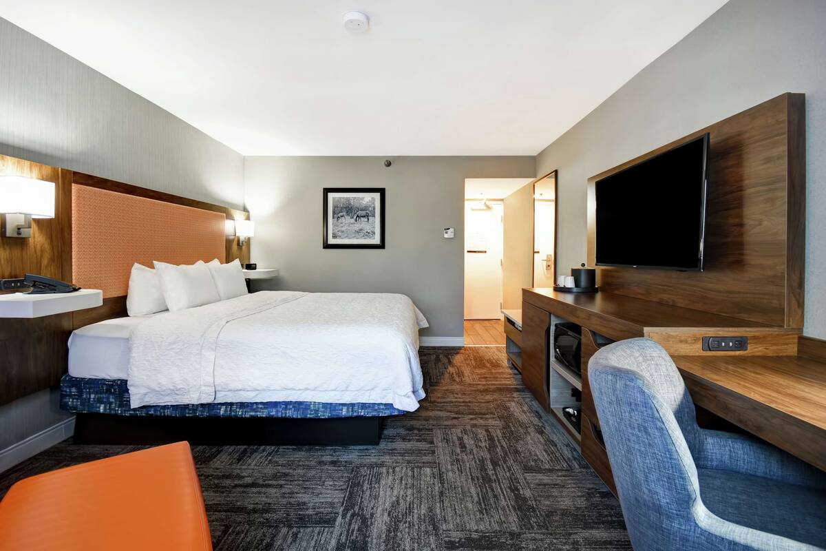 A hotel room with a bed, a desk with a flat-screen TV, a chair, and a nightstand.
