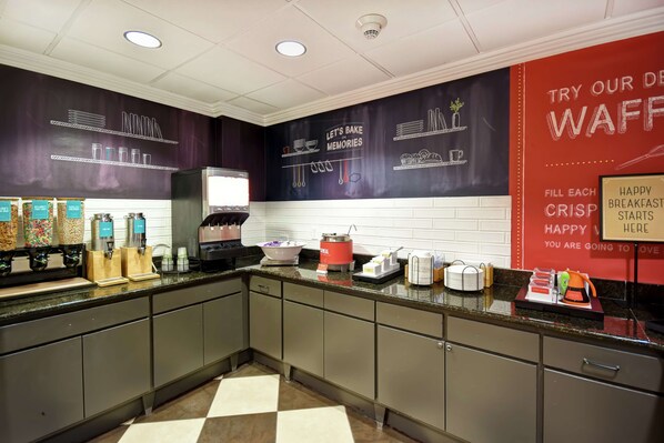 Un área de cocina con una estación de café, una máquina para gofres y un cartel que anuncia gofres.