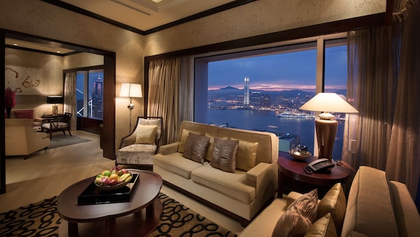 A hotel room with a large window offering a cityscape view, a sofa, a dining area, and a small table with fruit. - Conrad Hong Kong (Hong Kong)