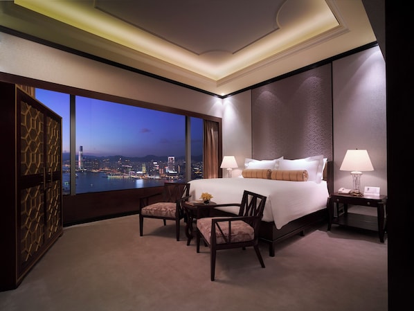 A hotel room with a large bed, two bedside tables, a desk, and a view of a cityscape at dusk.