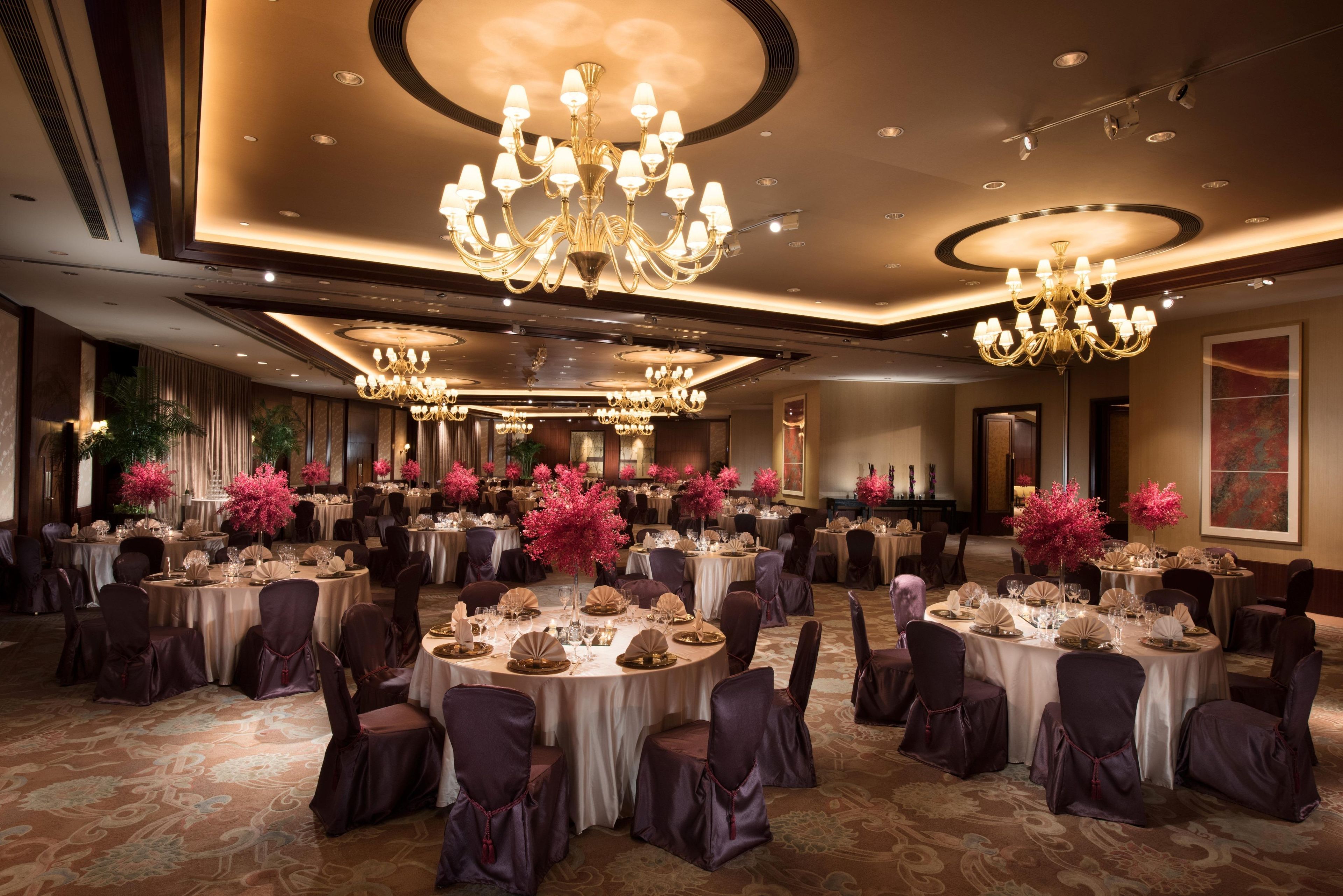 A banquet hall with round tables, chairs, chandeliers, and floral centerpieces.