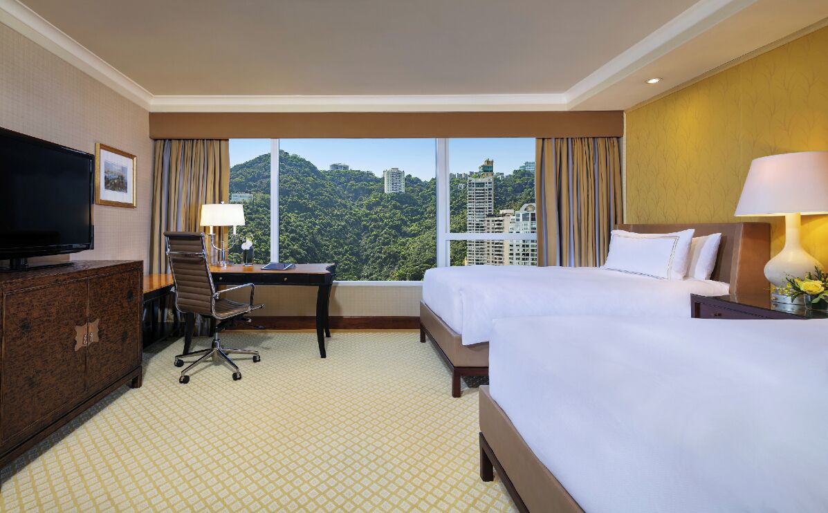 A hotel room with two beds, a desk, a TV, and a view of a cityscape.