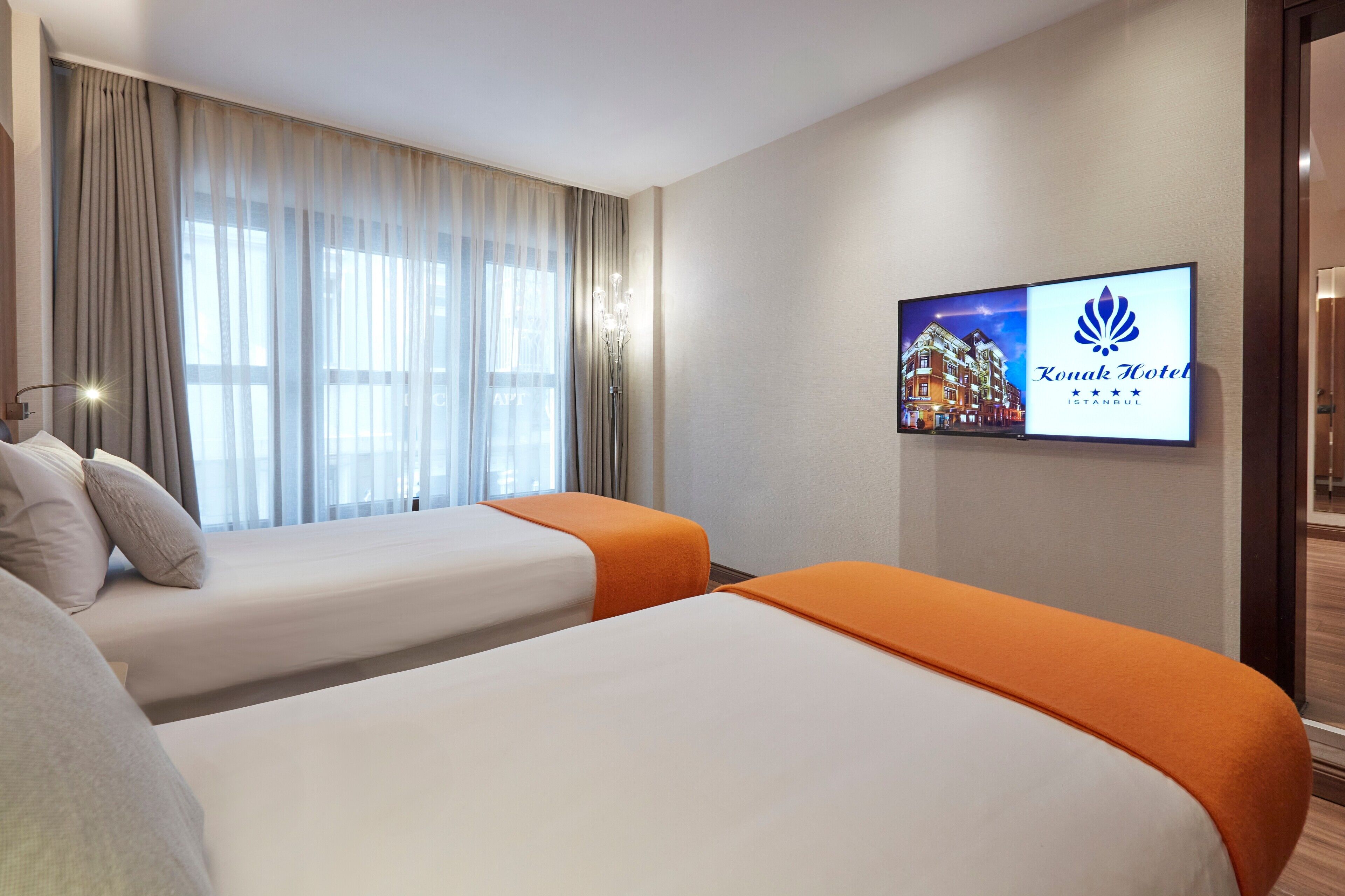 A hotel room with two beds, a TV displaying the hotel's name, and a window with curtains.