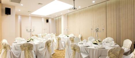 Un salón de banquetes con mesas redondas, manteles blancos y sillas con cintas doradas.