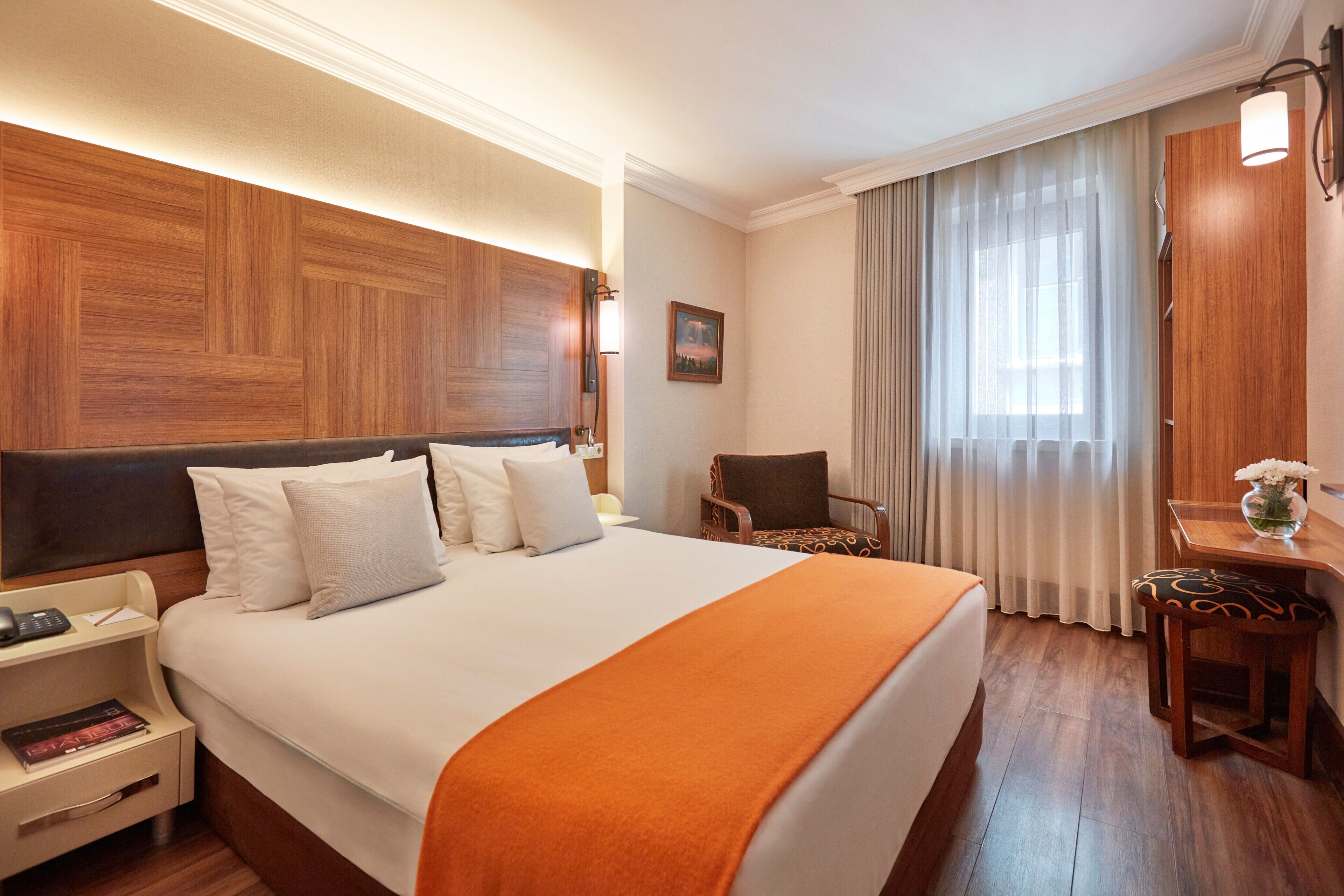 A hotel room with a large bed, a wooden headboard, a nightstand with a lamp, a chair, and a desk with a vase of flowers.