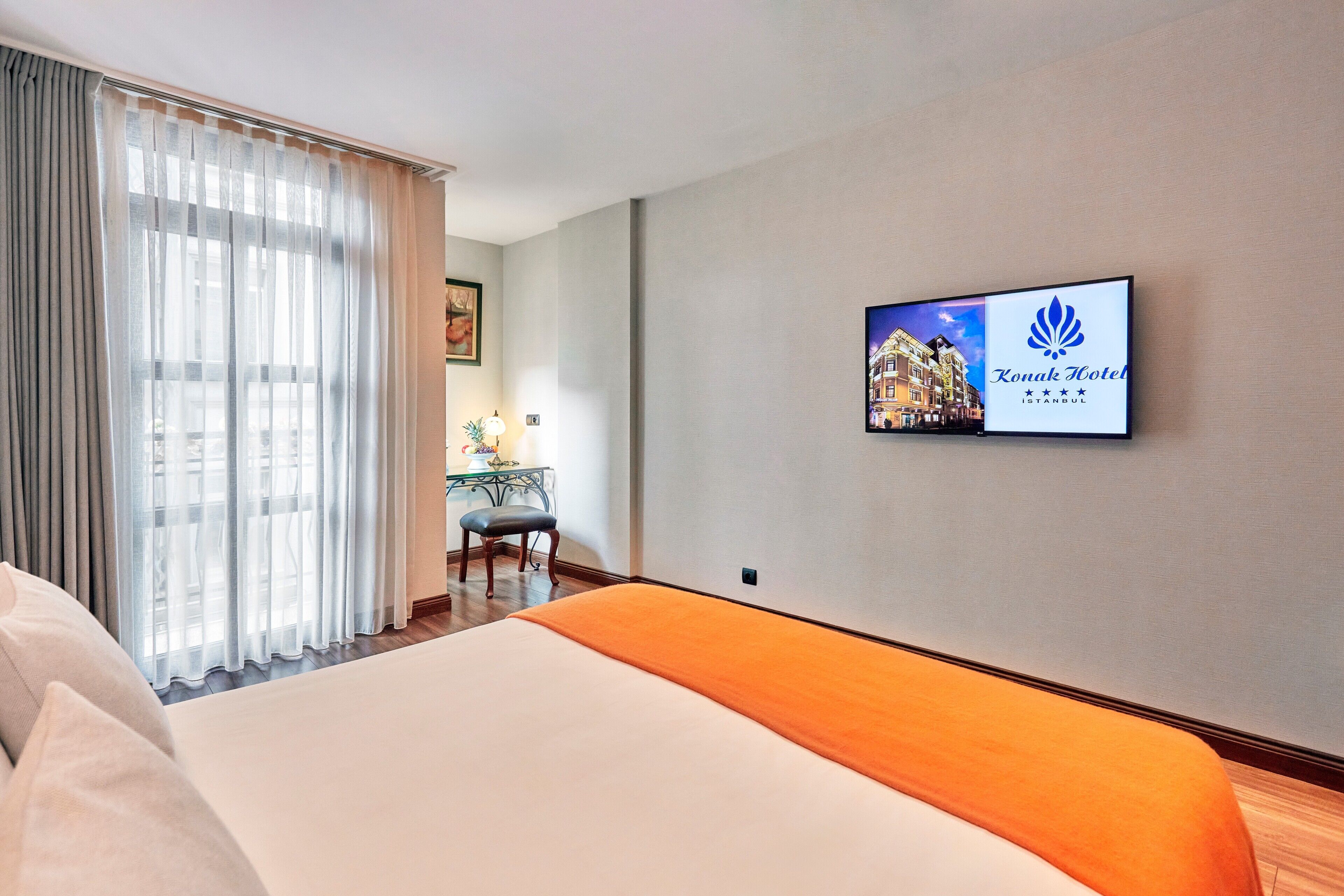 A hotel room with a bed, a desk with a chair, a window with curtains, and a wall-mounted picture of the hotel.