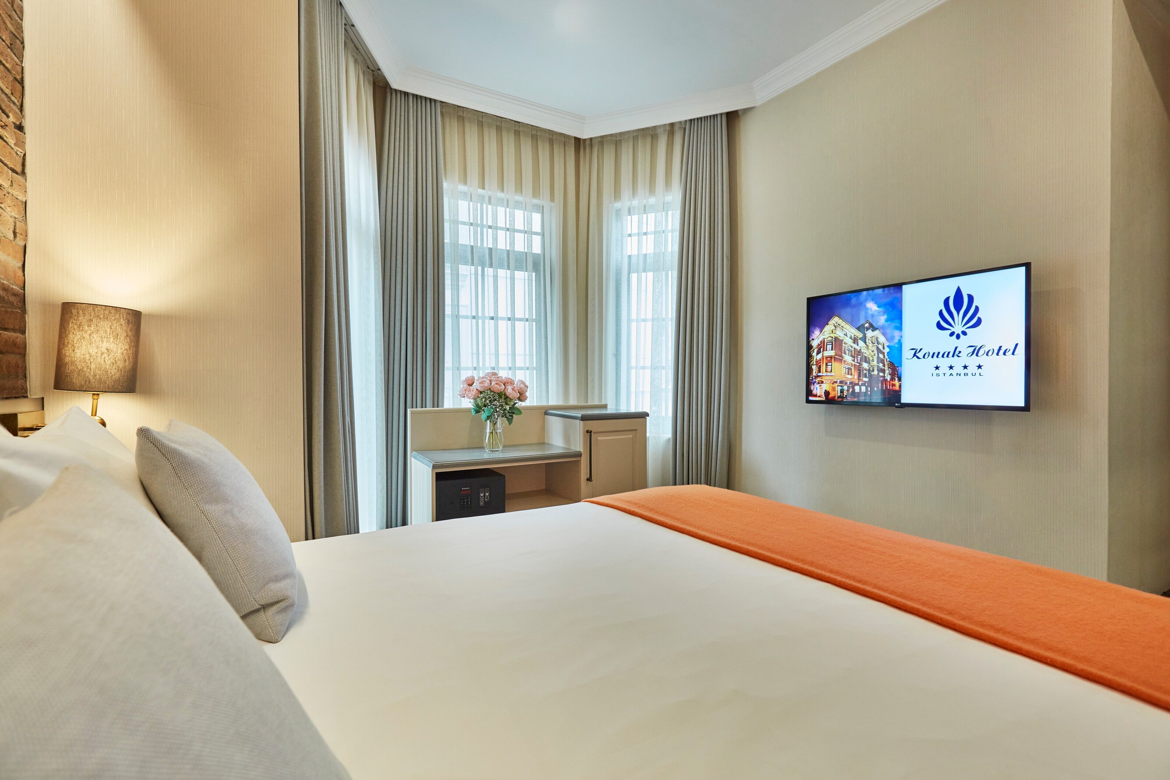 A hotel room with a bed, a desk, a TV displaying the hotel name, and a window with curtains.