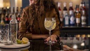 Un bar con una persona sosteniendo un vaso de una bebida cítrica, una botella y media lima sobre una mesa.