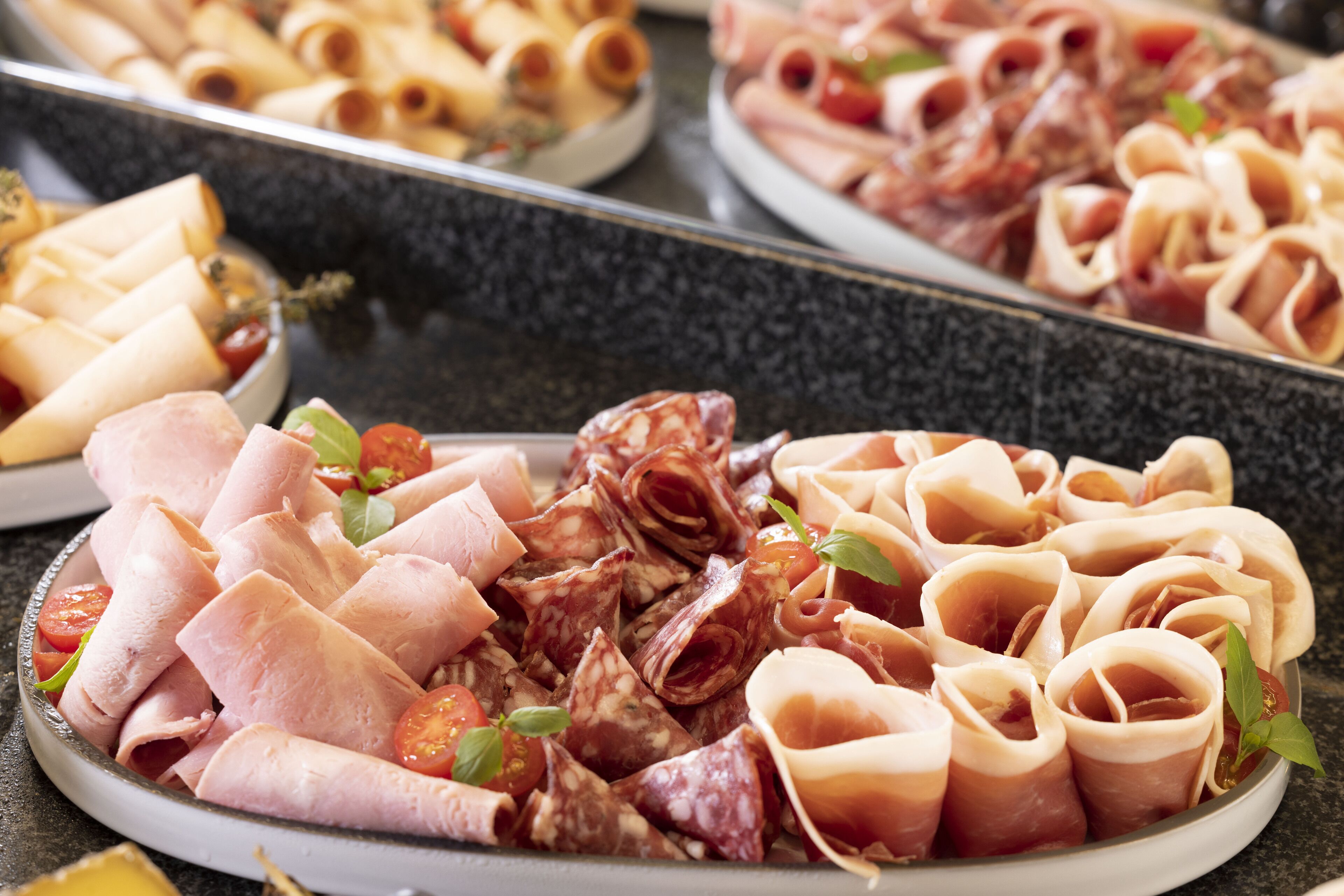 A platter of assorted meats and cheeses, including prosciutto, salami, and sliced ham, garnished with cherry tomatoes and basil leaves.
