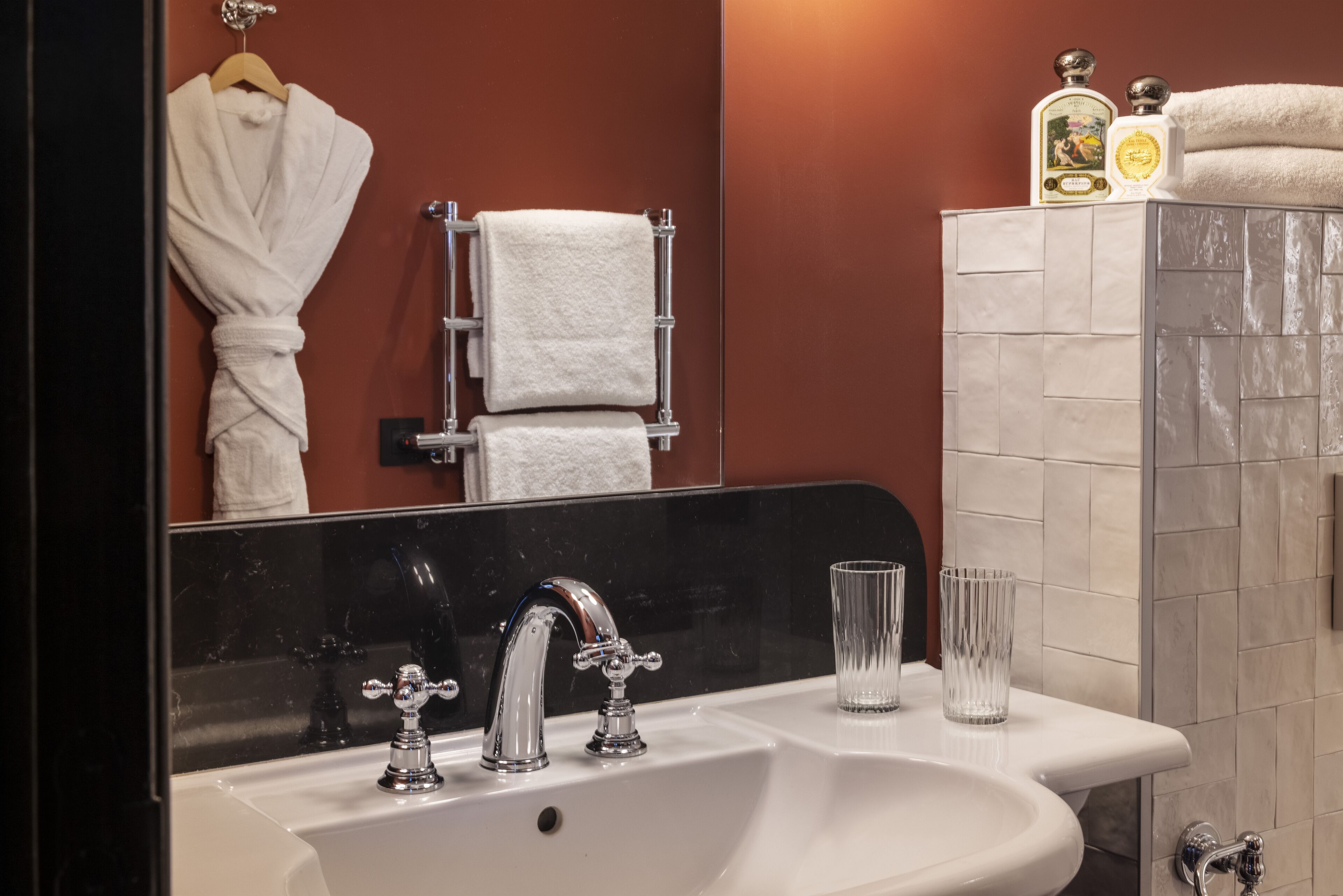 A bathroom with a white sink, two faucets, two clear glasses, a towel rack with two towels, and two bottles of lotion on a shelf.