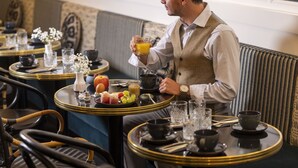 Un hombre sentado a una mesa con una bandeja de desayuno, que incluye frutas, un vaso de zumo de naranja y una taza de café.