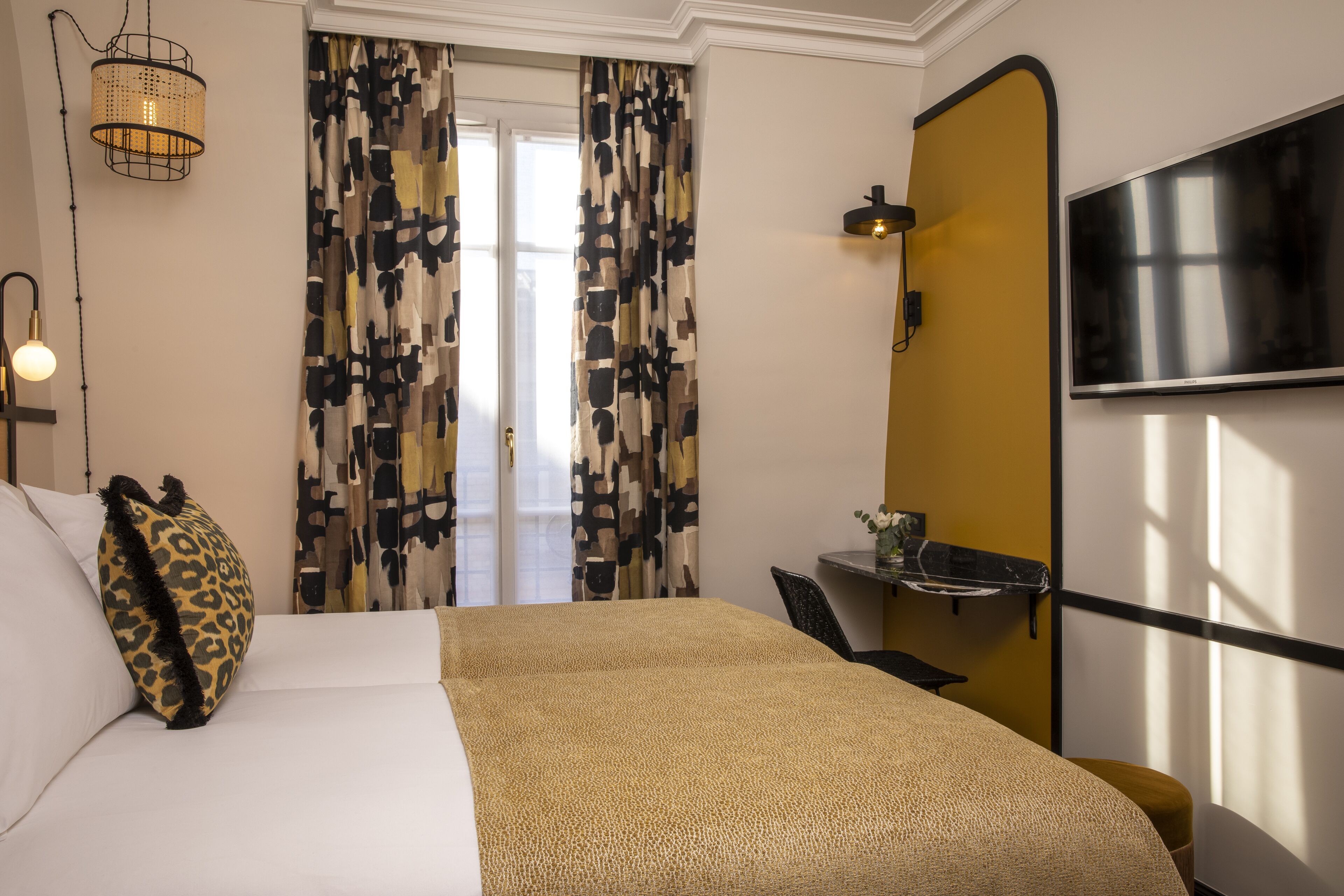 A hotel room with a bed, a desk, a television, and a window with patterned curtains.