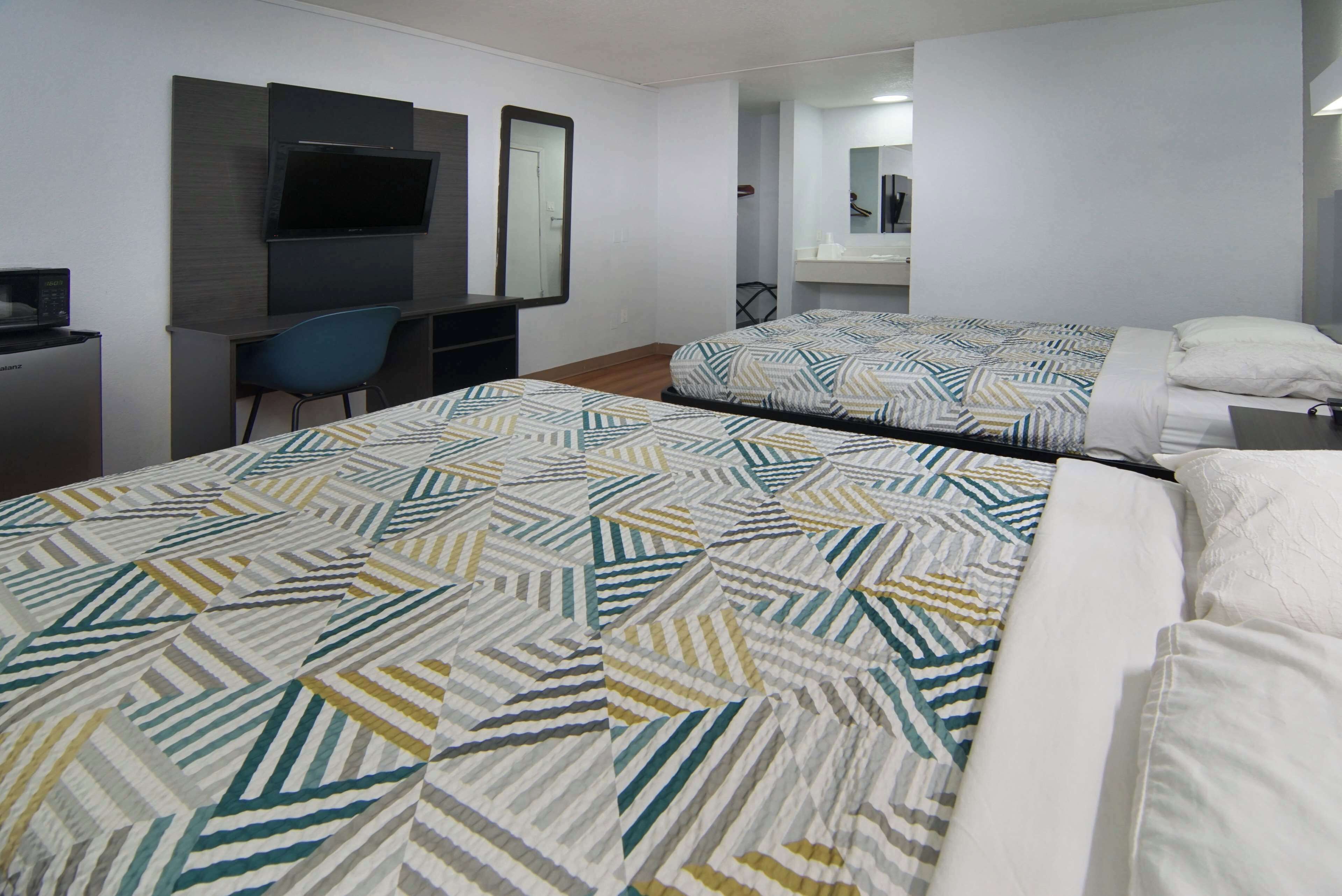 Ein Hotelzimmer mit zwei Betten, einem Schreibtisch mit Stuhl, einem an der Wand befestigten Fernseher und einem durchgegangenen Badezimmer.