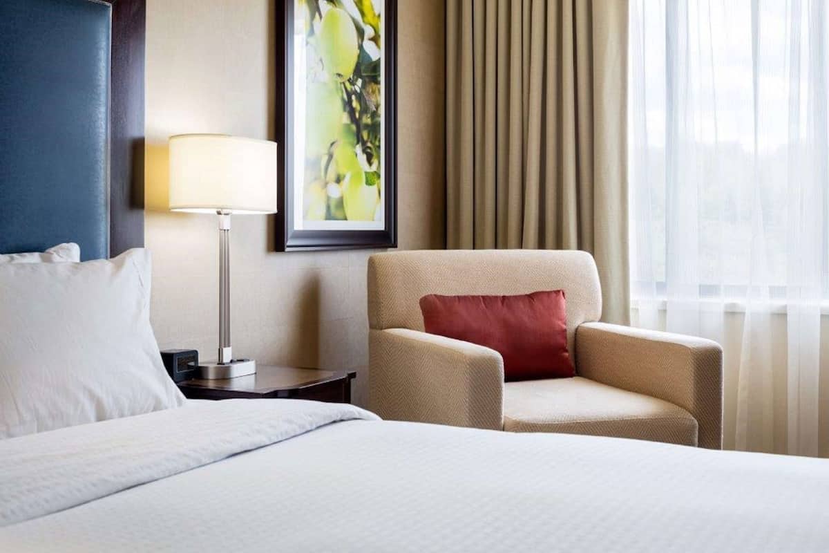 A hotel room with a bed, a chair, a nightstand, a lamp, a framed picture, and a window with curtains.