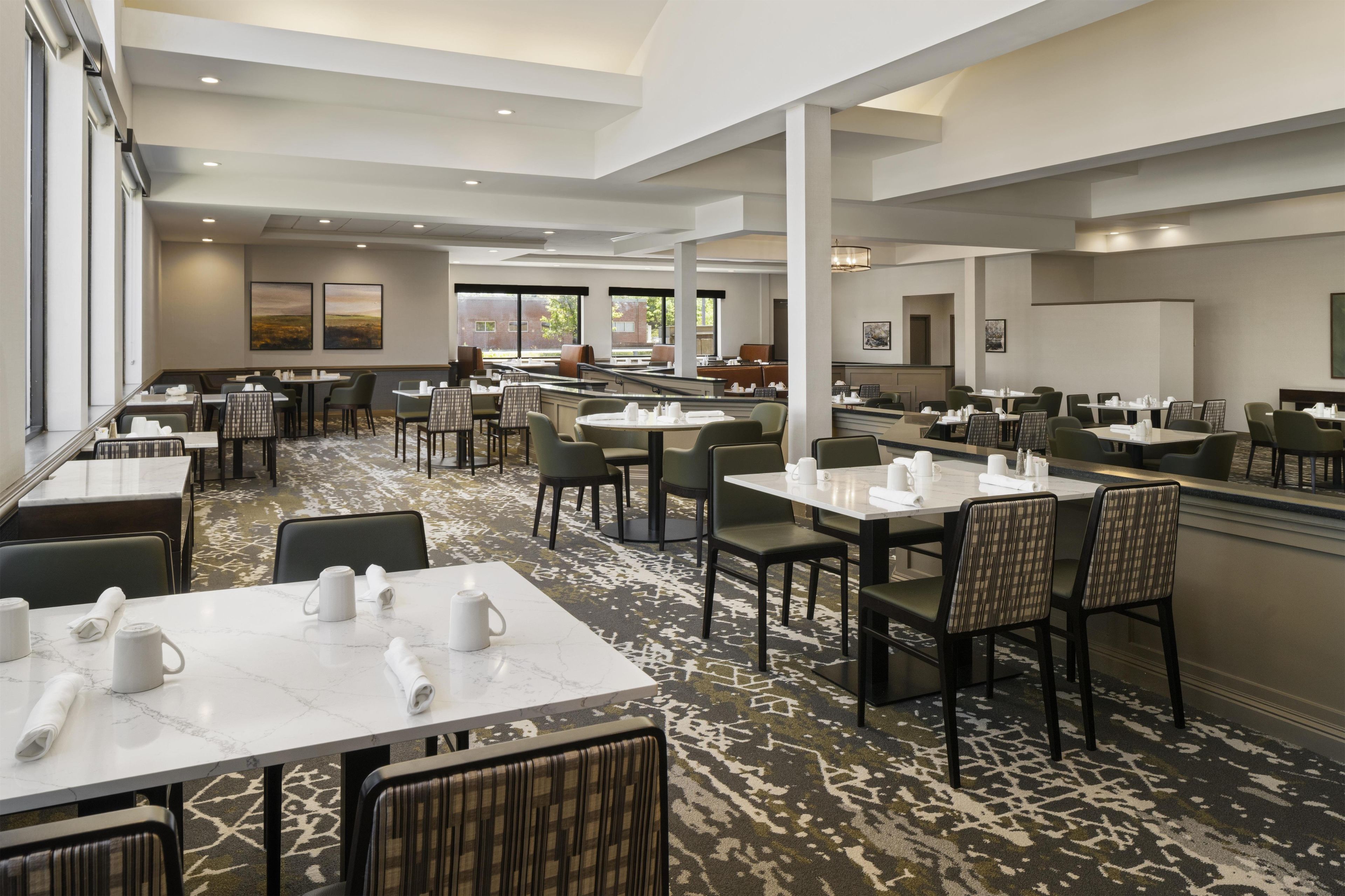 A spacious dining area with multiple tables and chairs, large windows, and artwork on the walls.