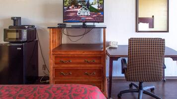 A hotel room with a television displaying 'Animal Cribs', a microwave, a wooden dresser with drawers, a patterned bedspread, and a chair.