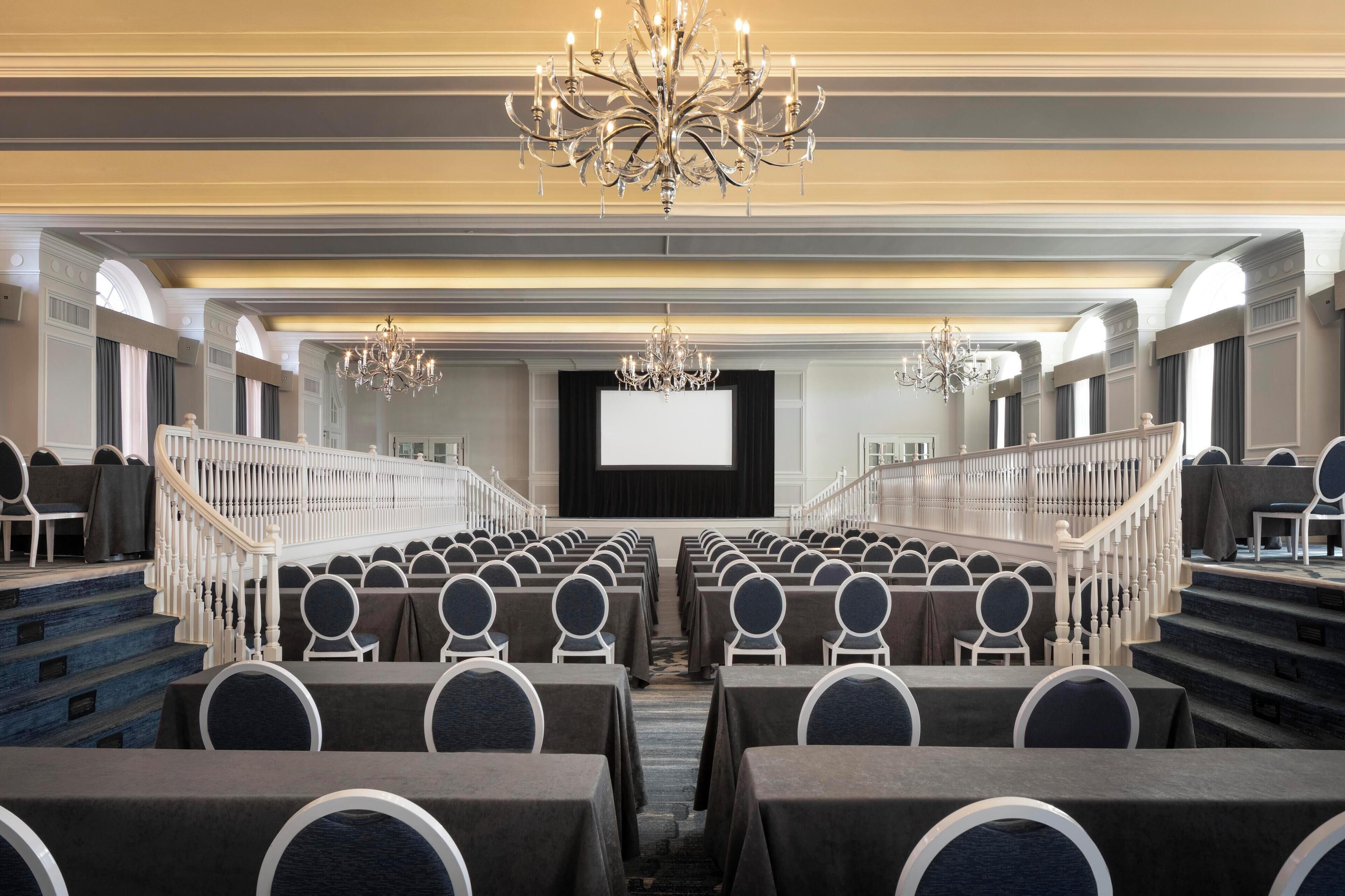 A conference room with rows of tables and chairs, a large screen at the front, and chandeliers.