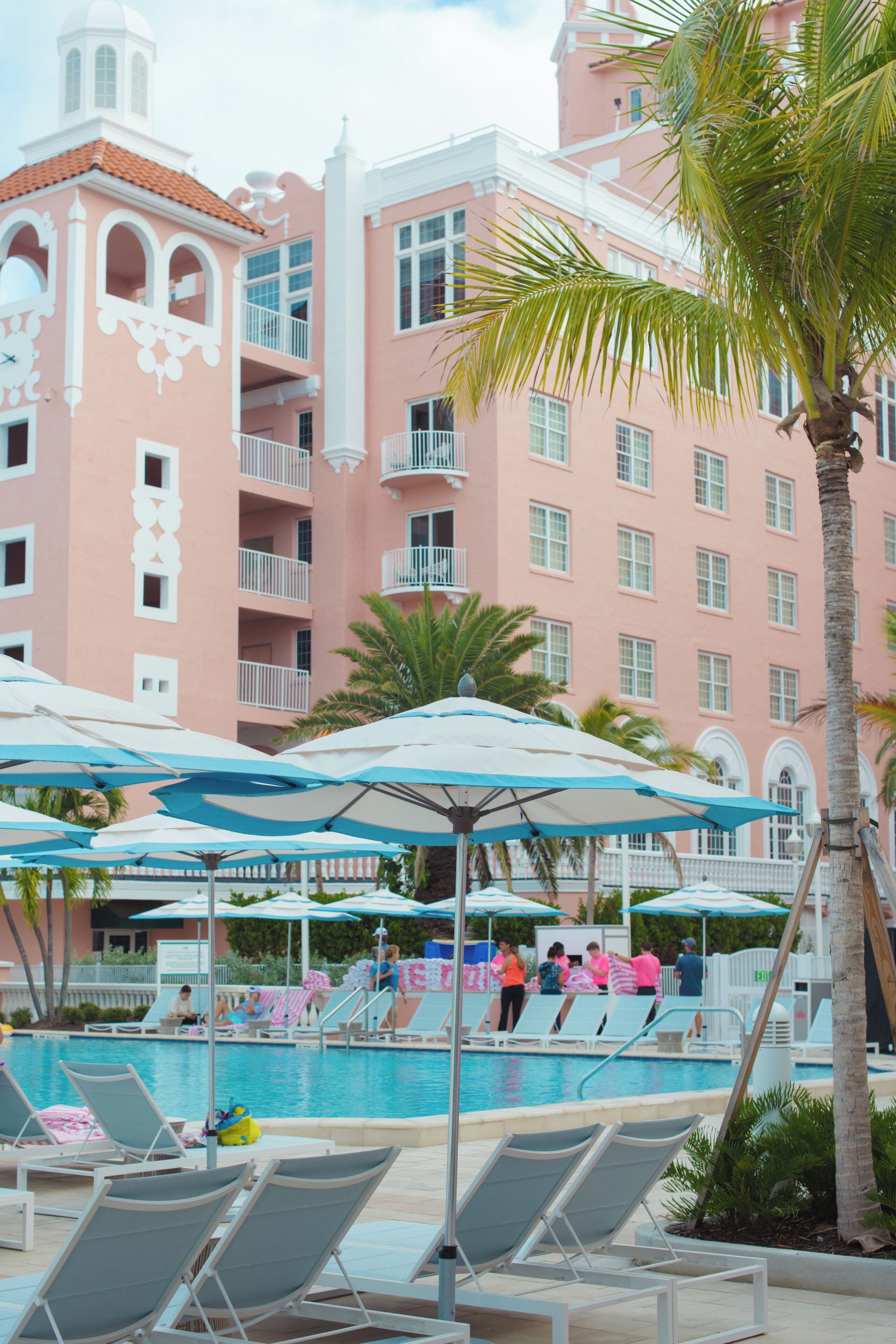 A pool area with lounge chairs and umbrellas, a pink building with balconies, and palm trees.