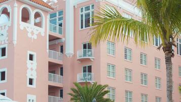 A pool area with lounge chairs and umbrellas, a pink building with balconies, and palm trees.