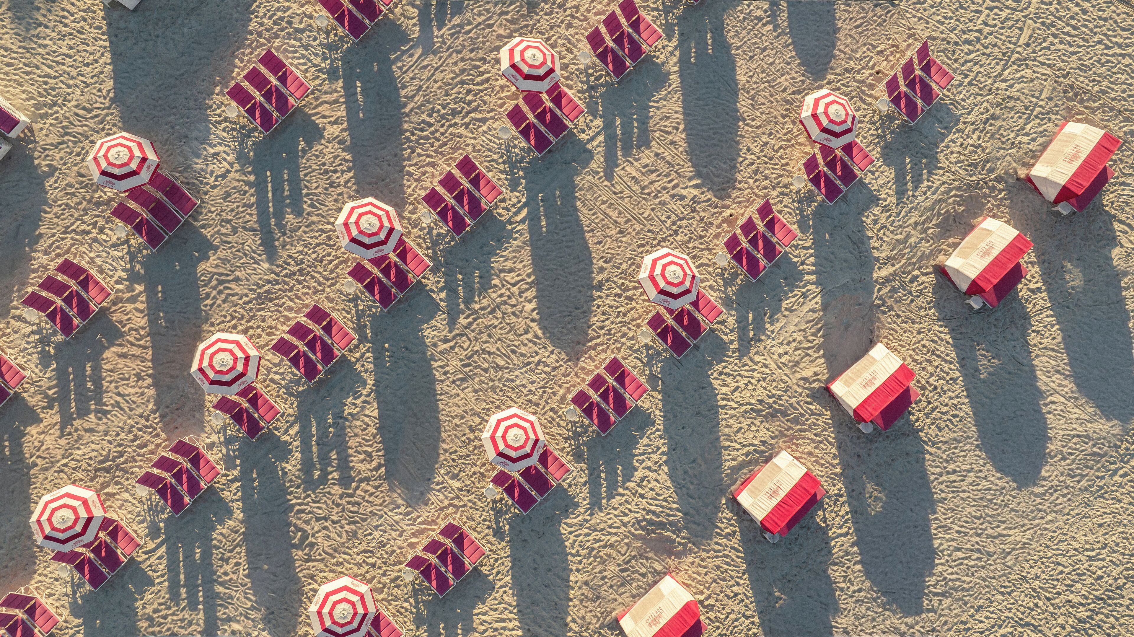 An aerial view of a beach with rows of red and white beach umbrellas and sun loungers arranged in a pattern.