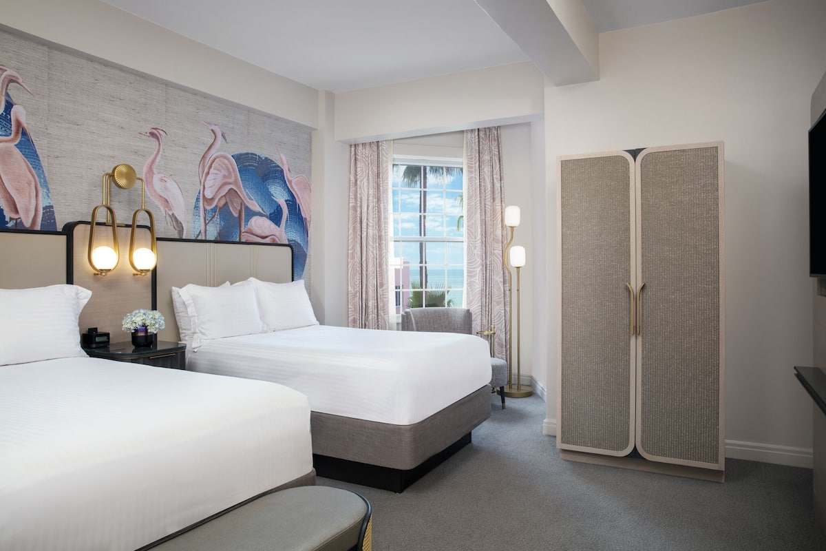 A hotel room with two beds, a large painting of birds on the wall, a view of the sea through a window, and a traditional wardrobe.