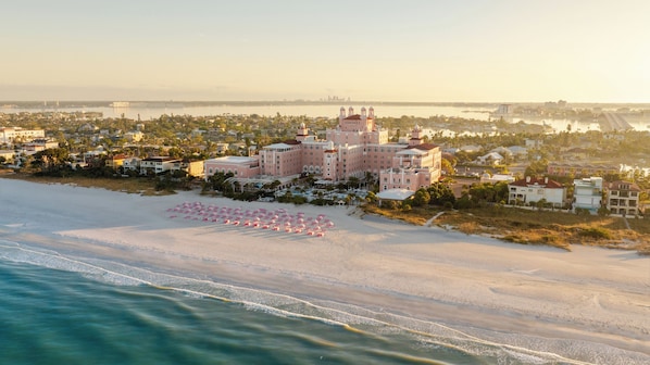 An aerial view of a coastal hotel with a beachfront, including a large building with multiple stories and a beach area with rows of pink umbrellas. - The Don CeSar (St. Pete Beach)