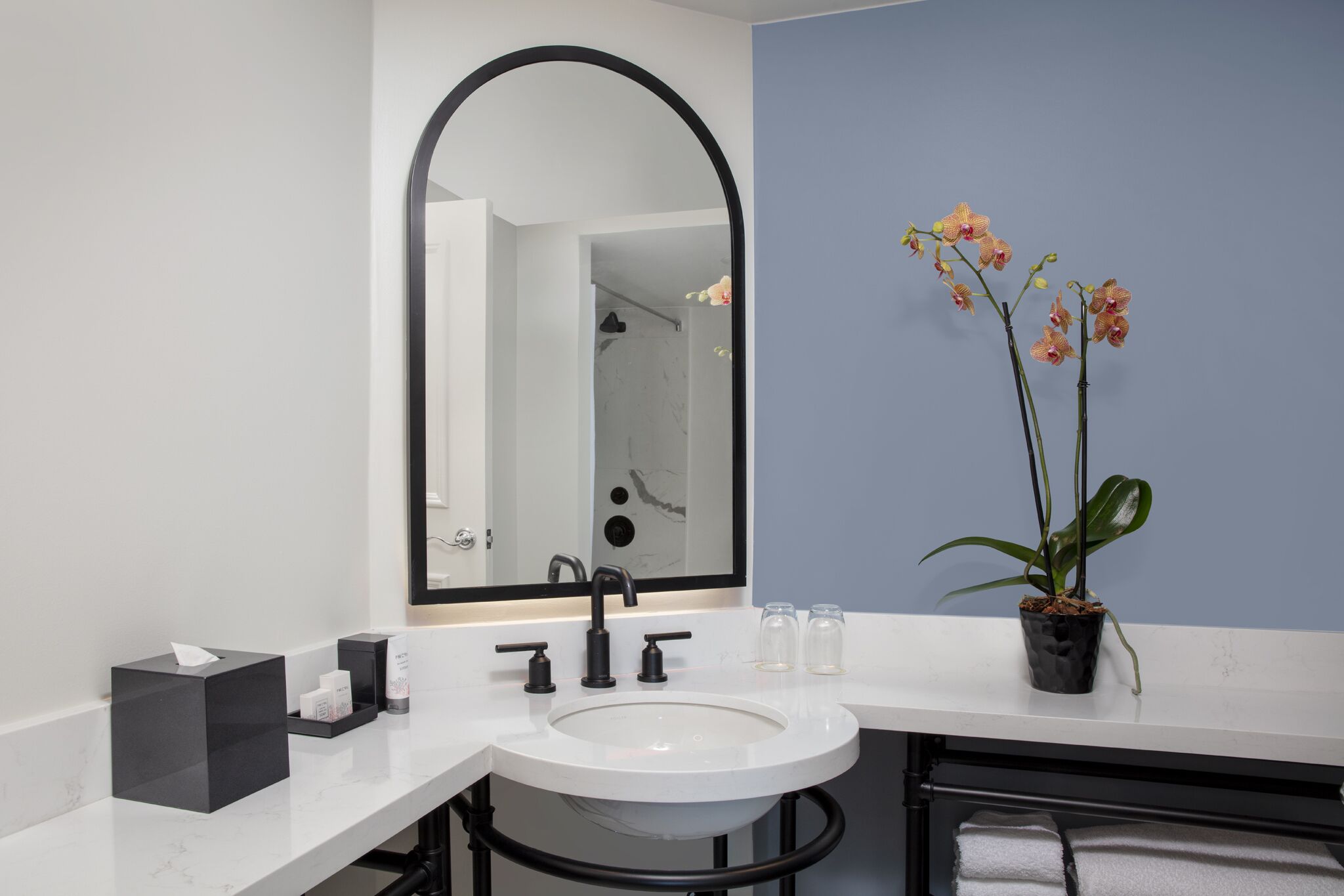 Un baño moderno con un lavamanos blanco, grifo negro y un espejo grande. Hay una orquídea en maceta sobre el mostrador y un perchero con una toalla.