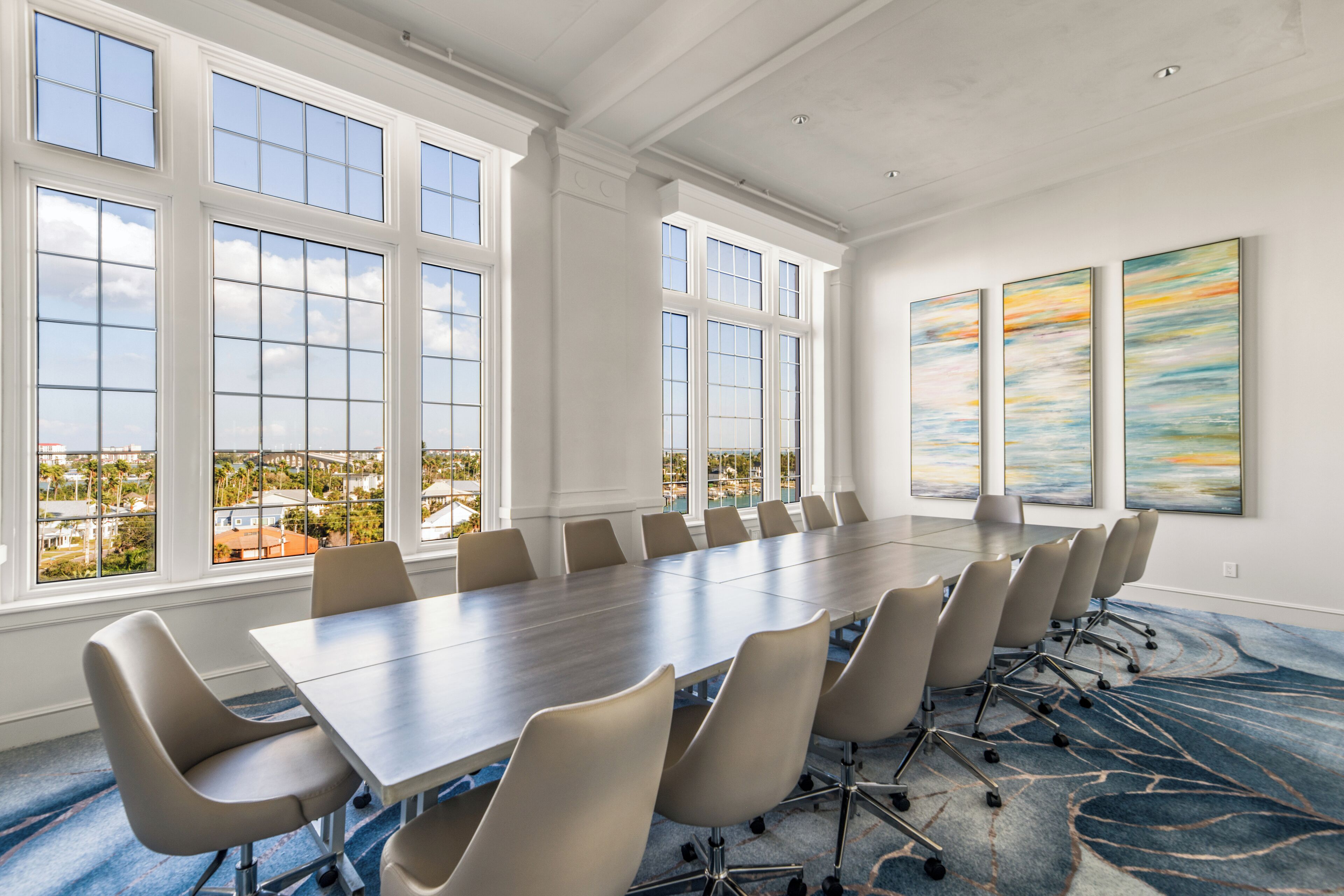 A conference room with a long table and chairs, large windows, and abstract paintings on the wall.