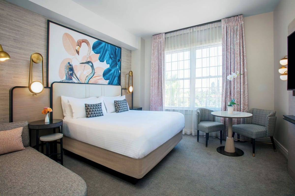 A hotel room with a large bed, a small round table, two chairs, a TV, and a painting on the wall.